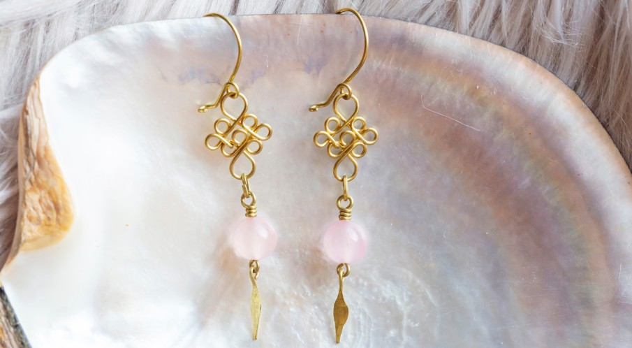A pair of gold wire statement earrings resting on a natural white shell background. The earrings have a pink mineral stone.