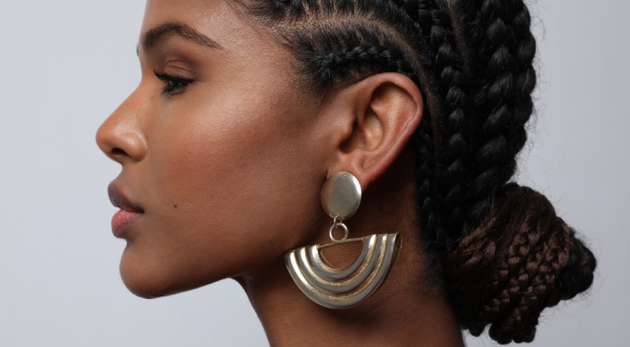 A profile view of a young woman wearing a pair of silver geometric statement earrings. Her hair is in a tightly braided bun.