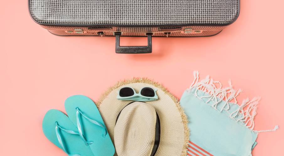 A pair of flip flops, sunglasses, straw hat, and beach towel are positioned below a closed suitcase.