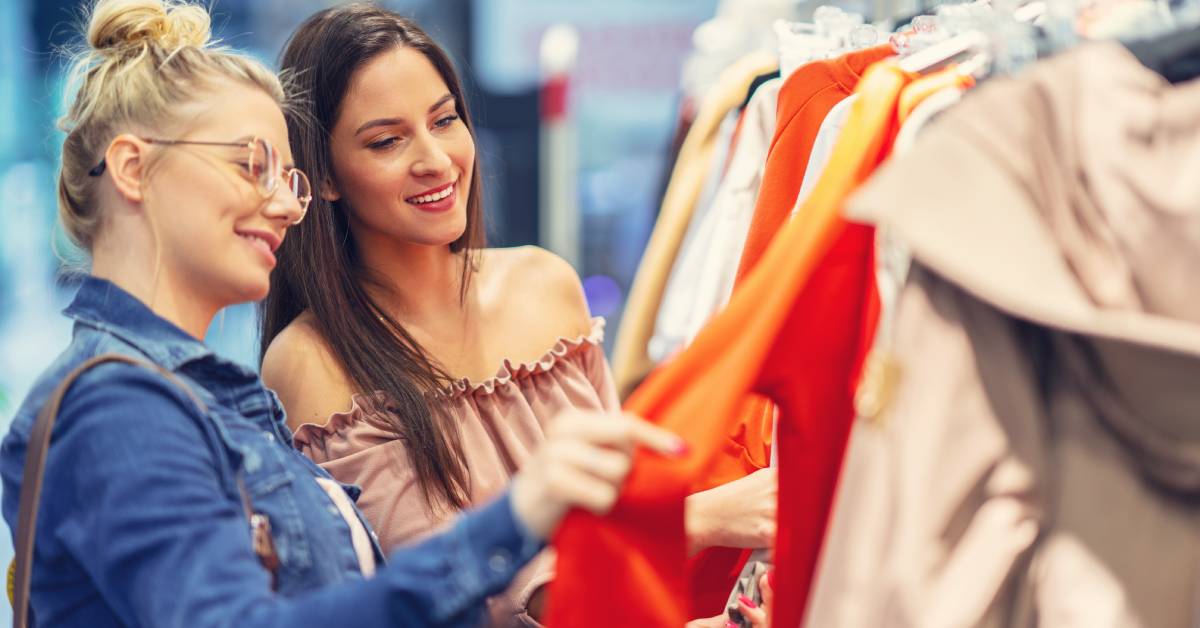 Two females are shopping together in a clothing store. They are smiling and looking at a rack of hanging clothes.