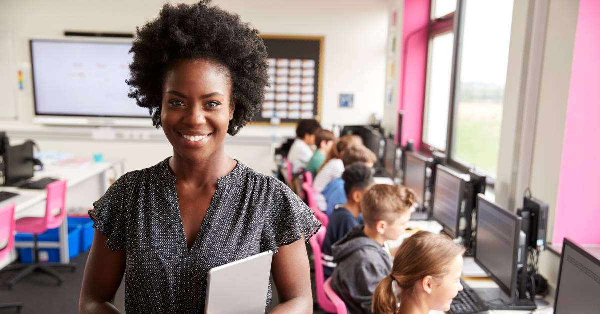 A female teacher is standing inside a computer lab with students. She is holding a tablet and wearing a lightweight dress.