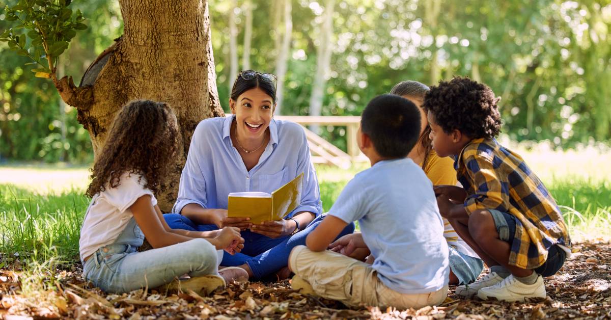 A female teacher and a group of four students are sitting outside in a circle. The teacher is reading a book to the students.