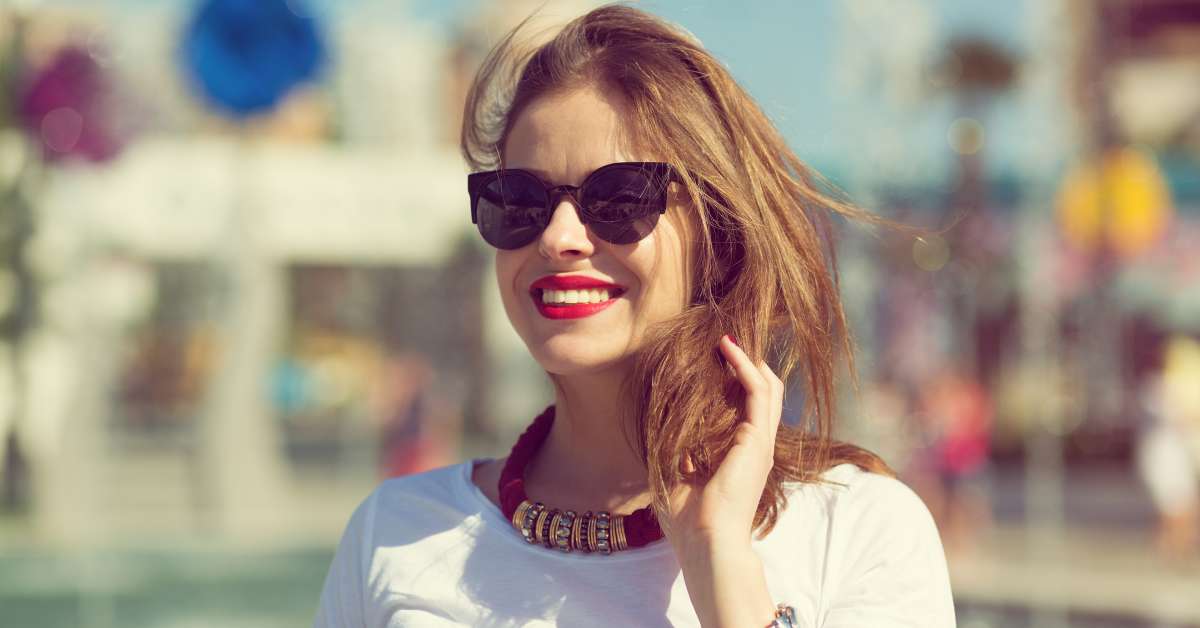 A woman smiling and touching her hair. She is wearing a t-shirt, a watch, a statement necklace, and a pair of sunglasses.