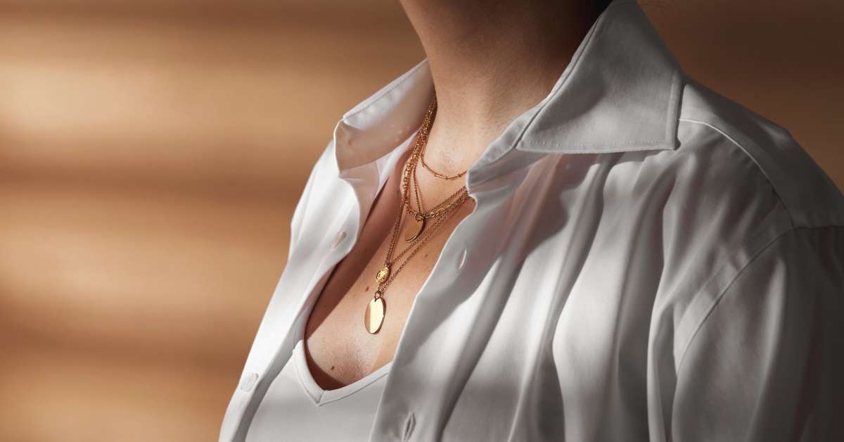 A woman wearing a white button-down shirt and several layered gold necklaces. Some of the necklaces have medallions.