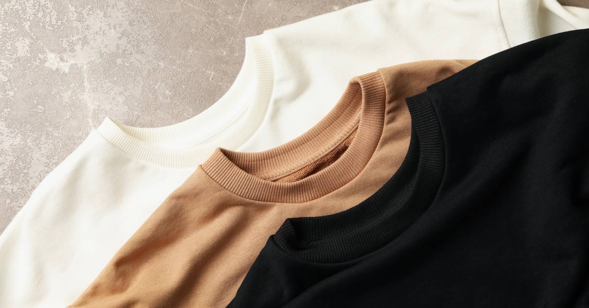 Three neutral crewneck t-shirts neatly lying on top of one another. One shirt is white, one is beige, and one is black.