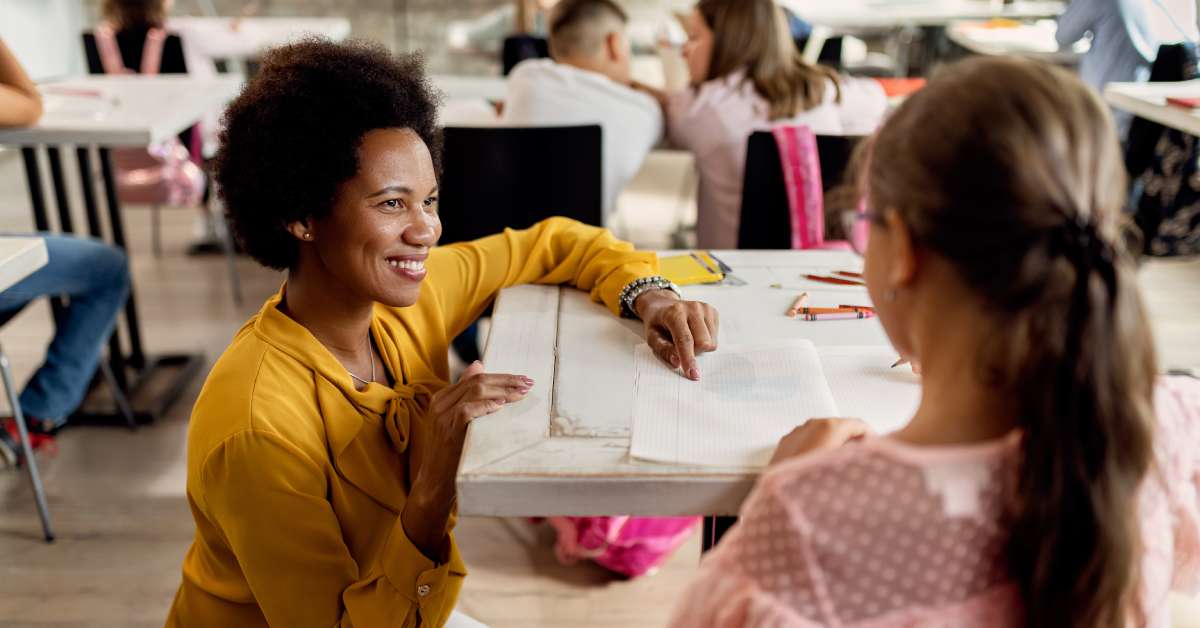 A smiling teacher is bending down to help a student sitting at a desk. She is wearing a yellow blouse and white pants.