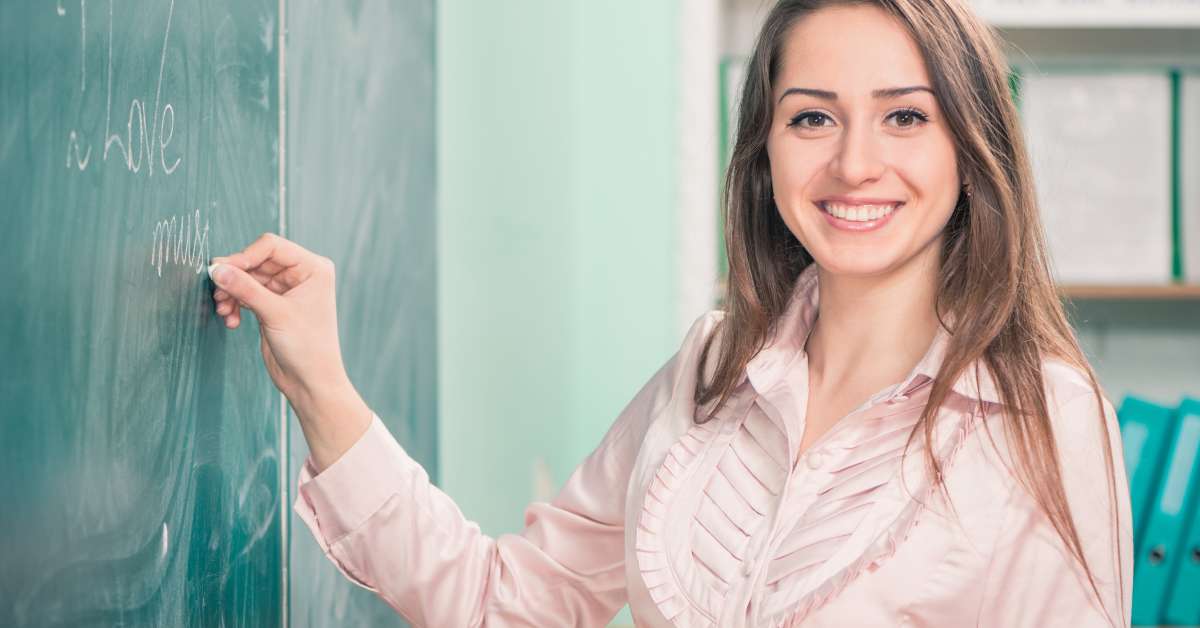 A smiling teacher is holding a piece of chalk and writing on a chalkboard. She is wearing a pink blouse with ruffles.