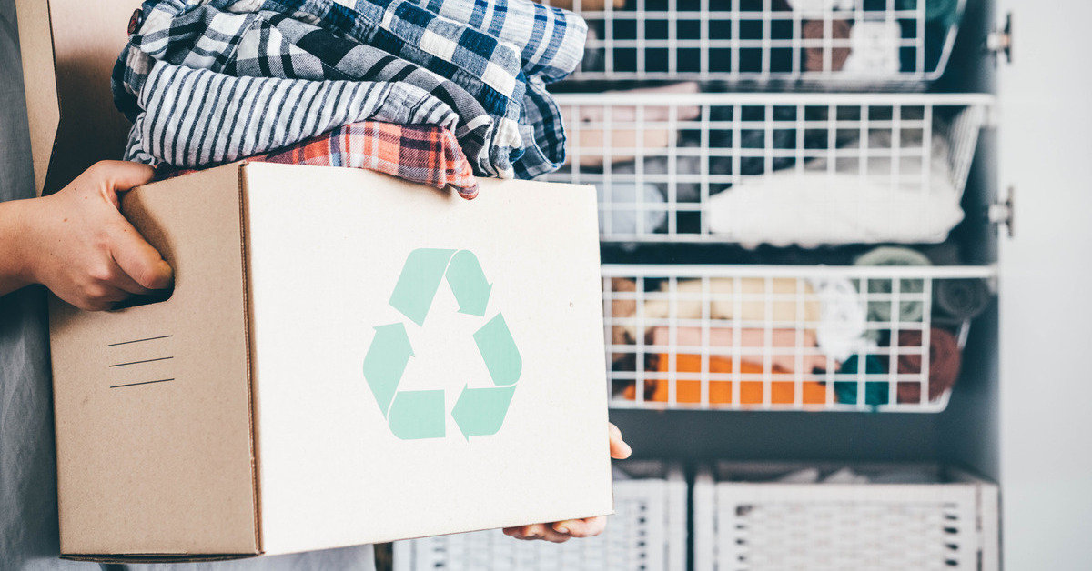 An individual is holding a brown box with a green recycling symbol on the front. The box is full of patterned clothing.