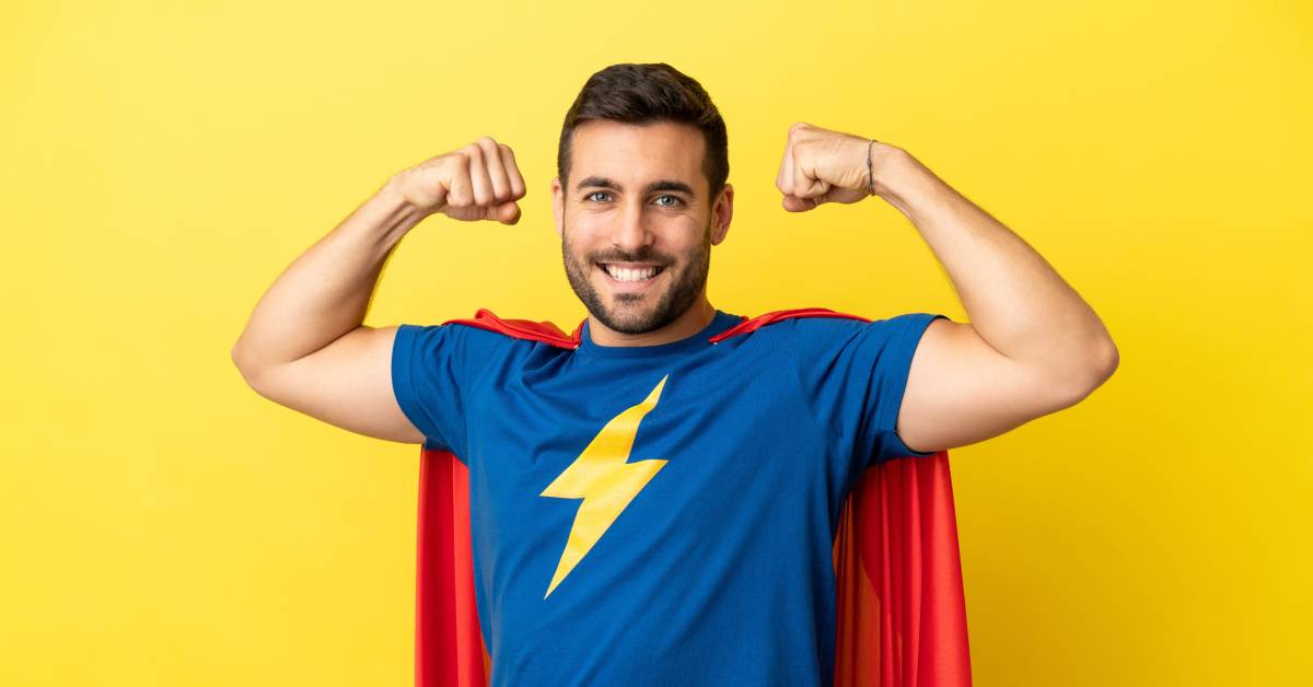 A man is wearing a blue T-shirt with a yellow lightning bolt on it and a red cape. He is smiling and flexing his muscles.